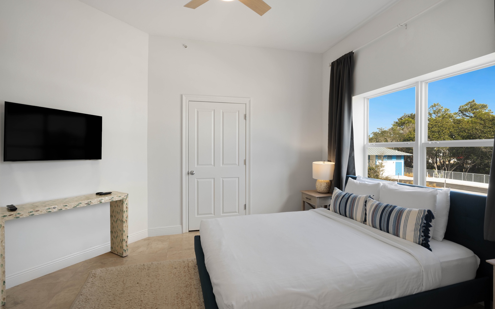 Unwind in your bright, airy bedroom featuring crisp linens, mounted TV, and peaceful tree views through large windows.