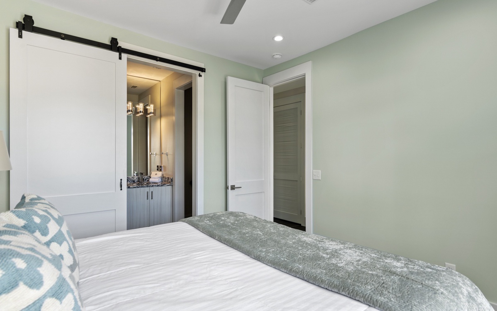 Your peaceful bedroom features soothing sage walls, crisp linens, and a unique barn-door bathroom entrance for modern coastal charm.