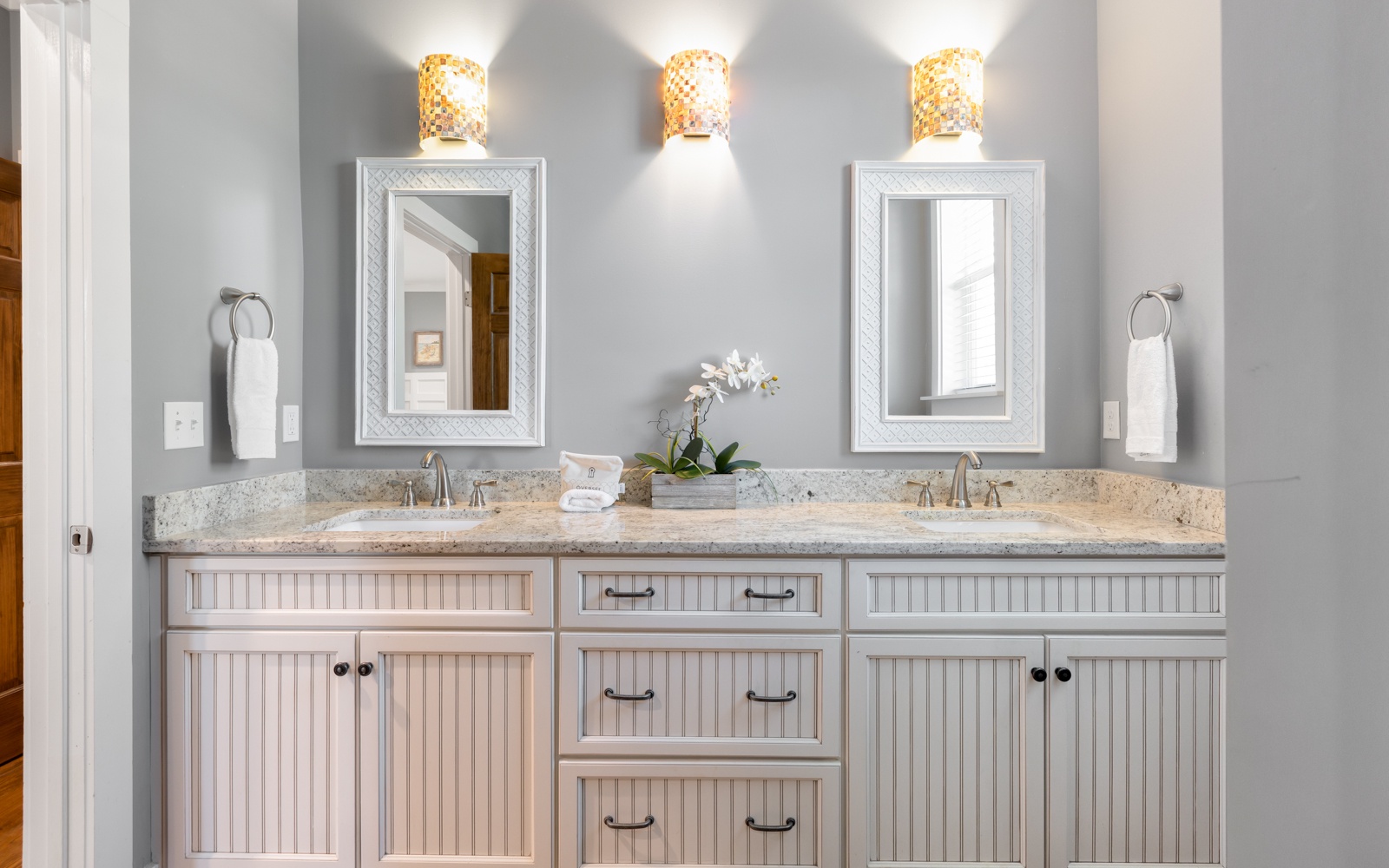Unwind in your elegant dual-sink bathroom featuring warm gold lighting, granite countertops, and thoughtful touches like fresh orchids.