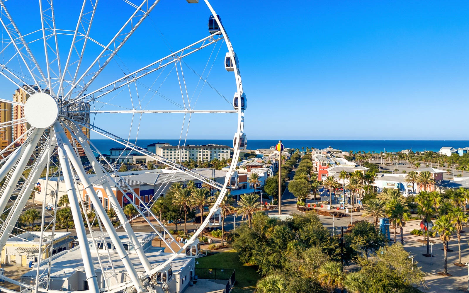 White ferris wheel overlooking a vibrant coastal town with palm trees, colorful buildings, and ocean views stretching to the horizon.