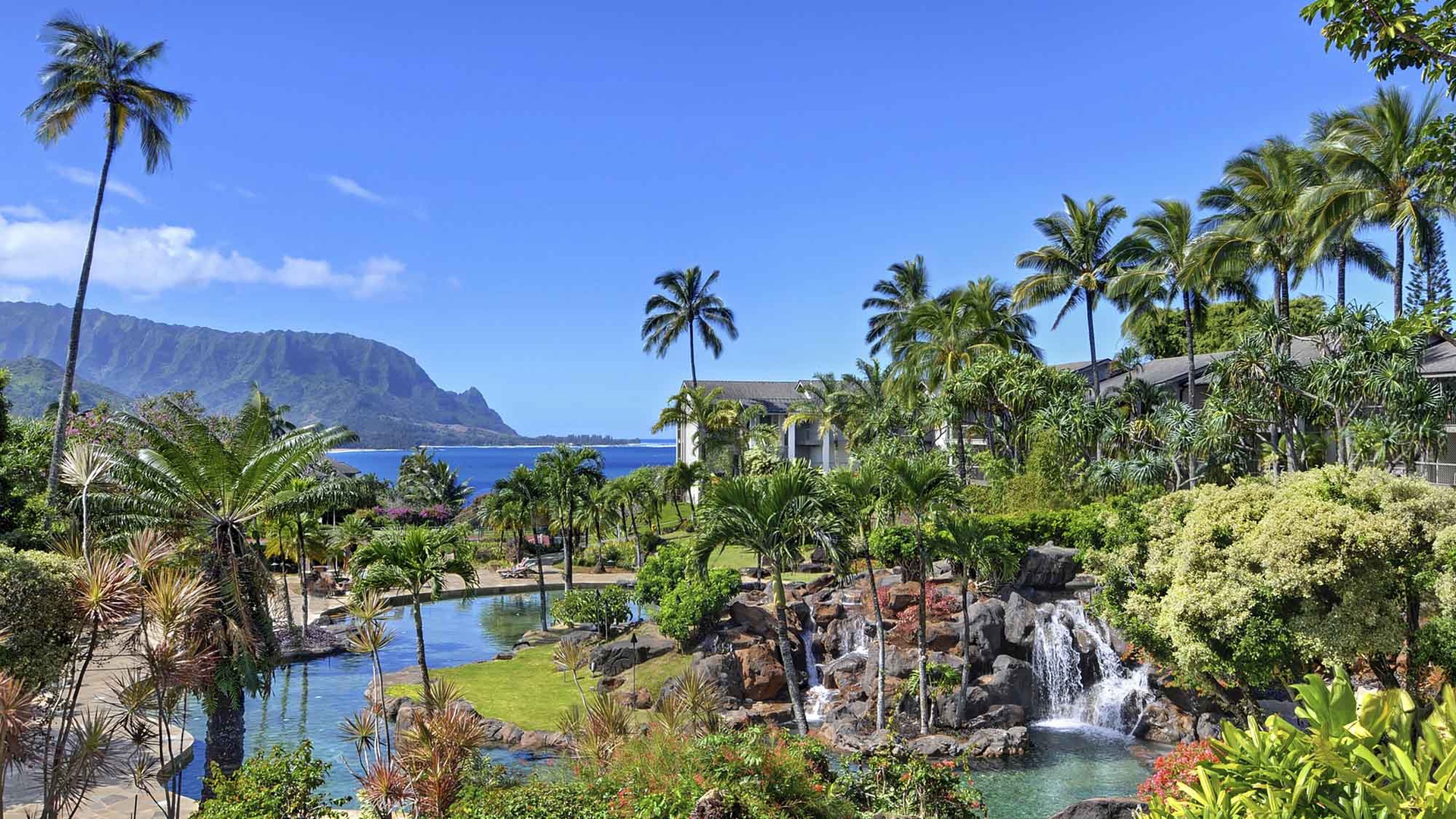 Tropical resort grounds featuring lush gardens, cascading waterfall, lagoon-style pools, and dramatic mountain and ocean views framing the property.