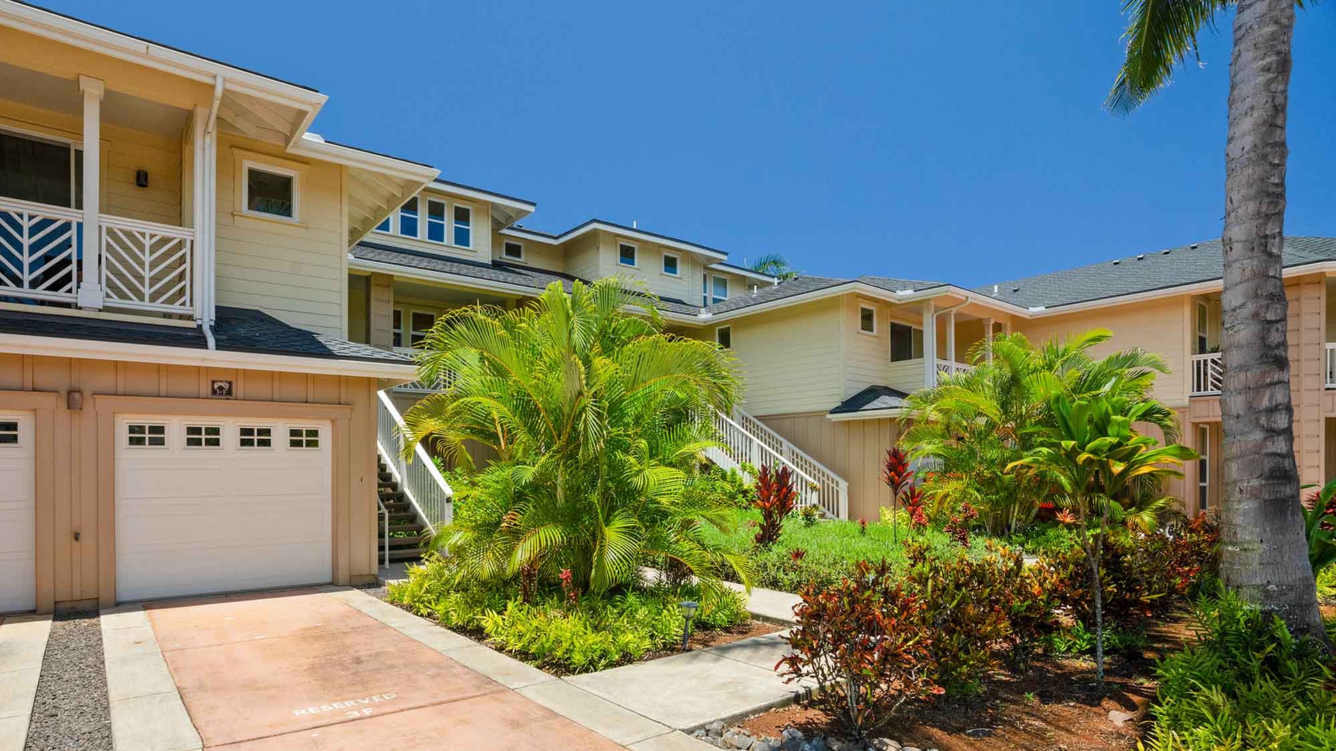 The property features a tropical island-style building surrounded by lush landscaping, including palm trees and vibrant foliage, with covered parking and exterior stairways leading to the units.