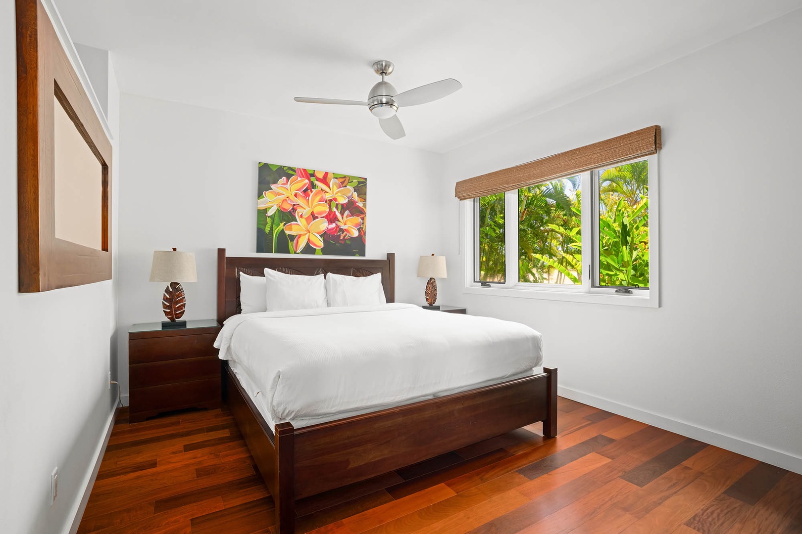 Unwind in your serene bedroom with rich hardwood floors, tropical garden views through picture windows, and a plush bed beneath a gentle ceiling fan to keep you cool and comfortable.