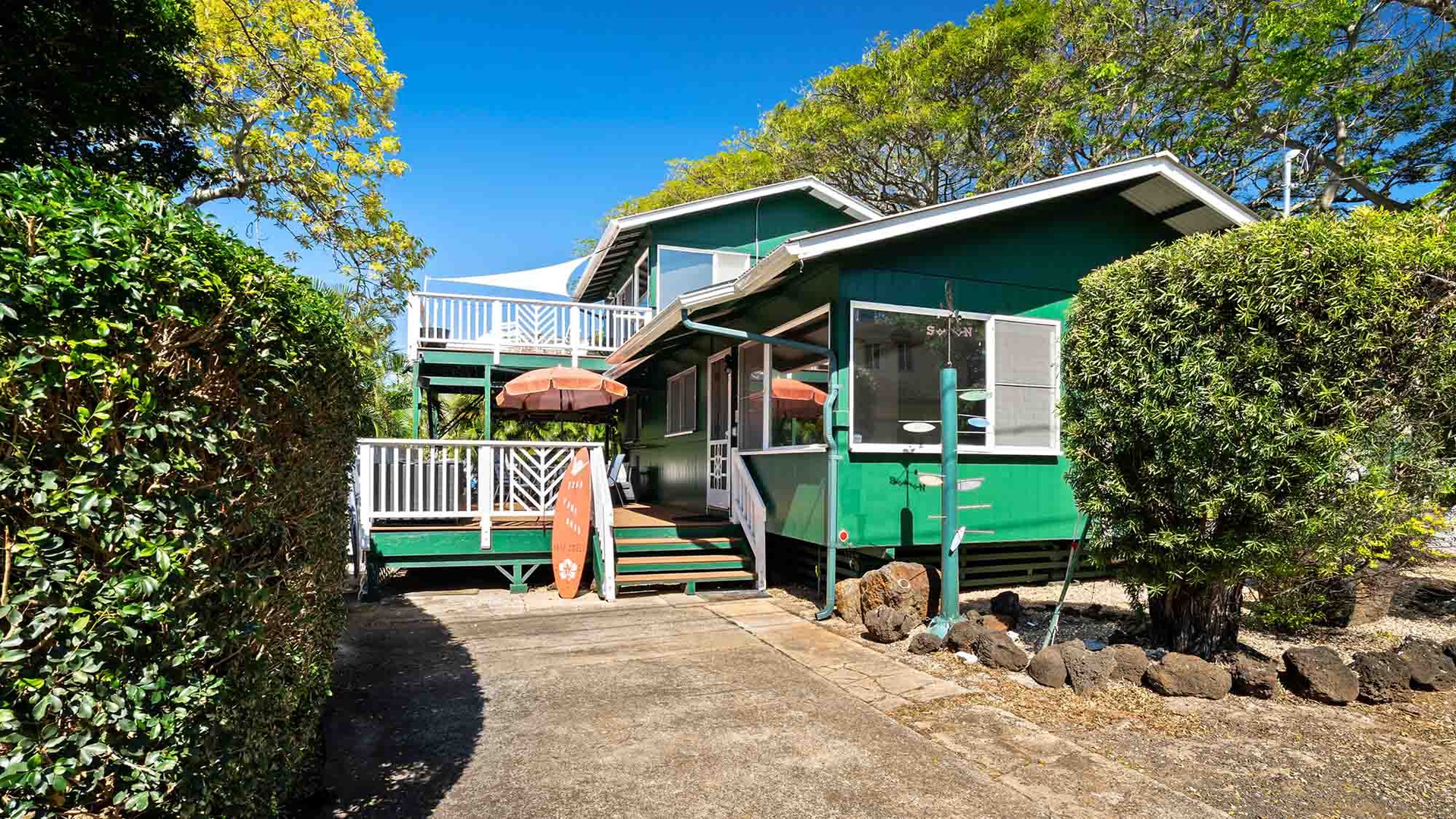 Charming green beach house with private deck and surfboard, nestled among tropical trees under brilliant blue skies.