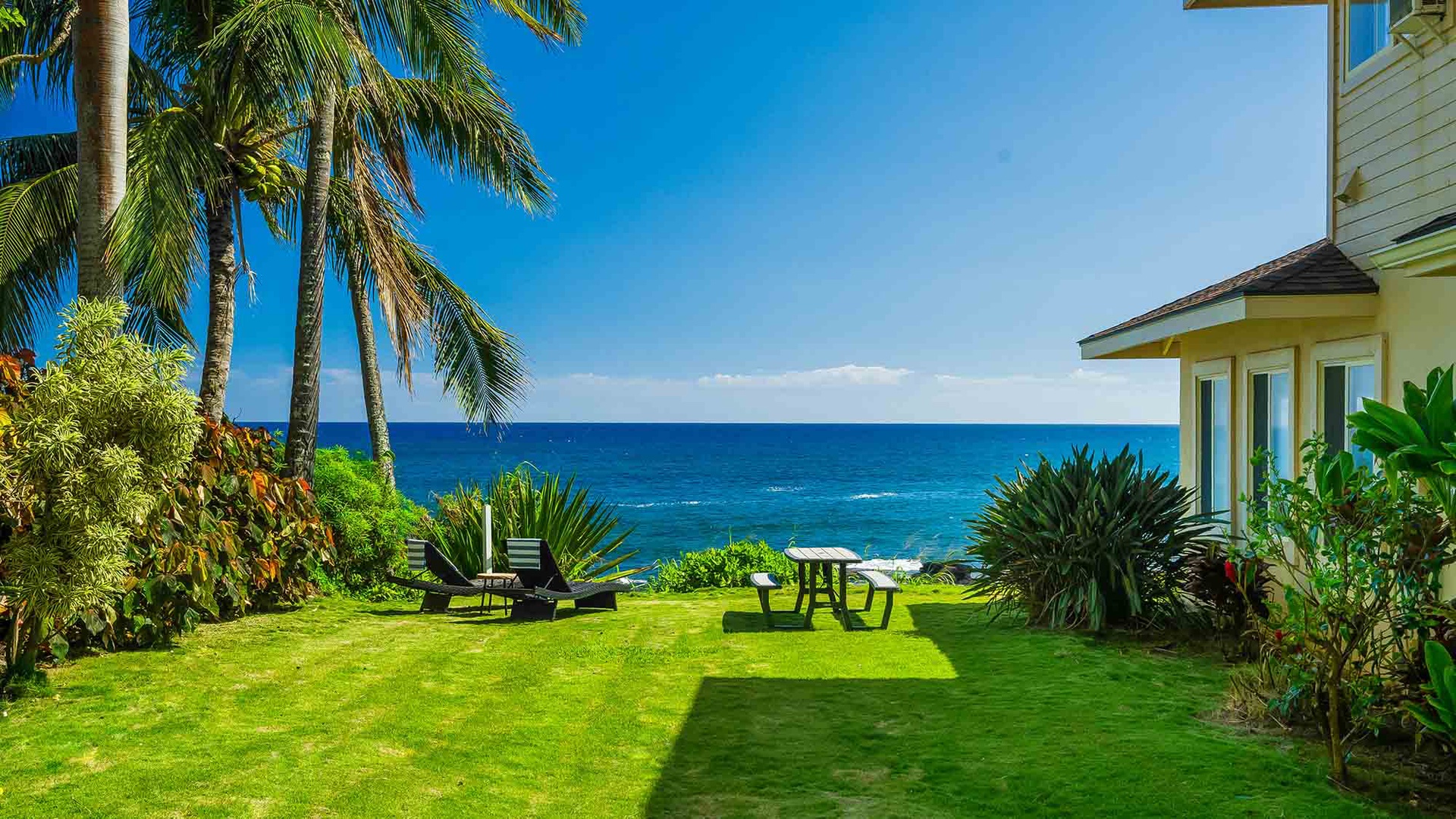 A beachfront property featuring a manicured lawn with tropical landscaping leading directly to the ocean, framed by palm trees.