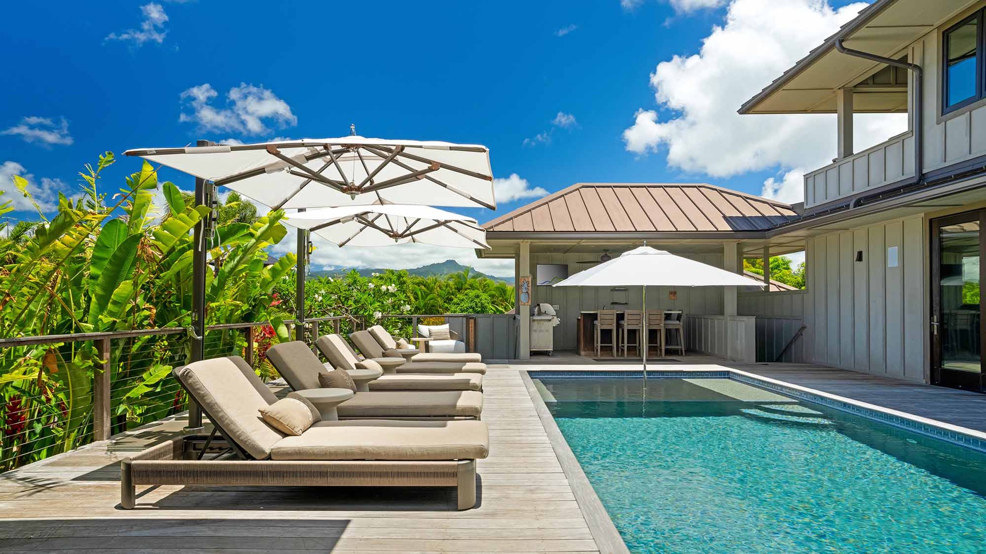 Stunning pool deck framed by lush tropical greenery and mountain views. Lounge on cushioned sunbeds or gather at the covered bar—your private retreat under bright island skies.