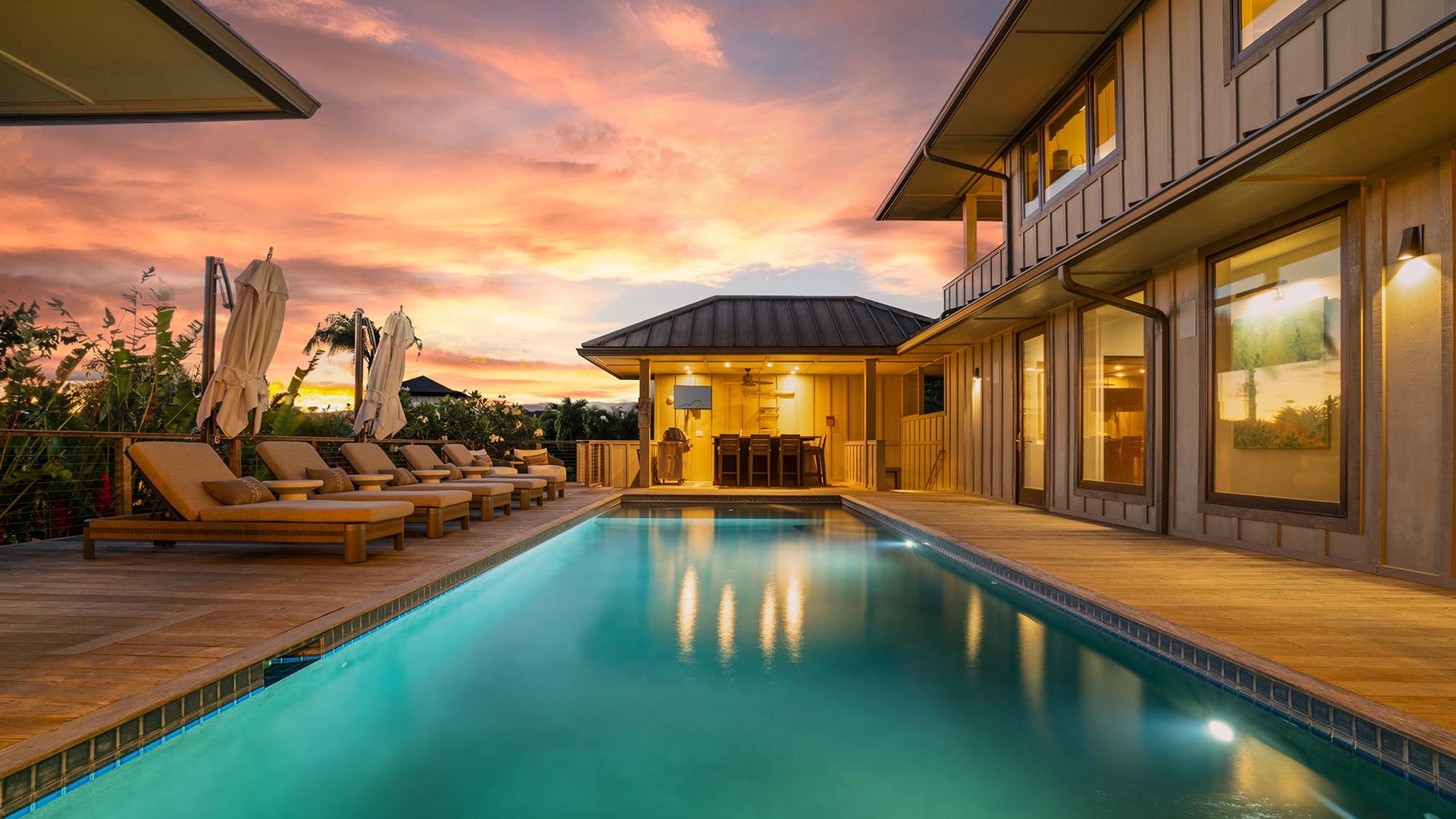 A stunning private villa features a pristine pool deck beneath a breathtaking sunset sky, with wooden loungers, tropical greenery, and warm interior lighting creating an idyllic island retreat.