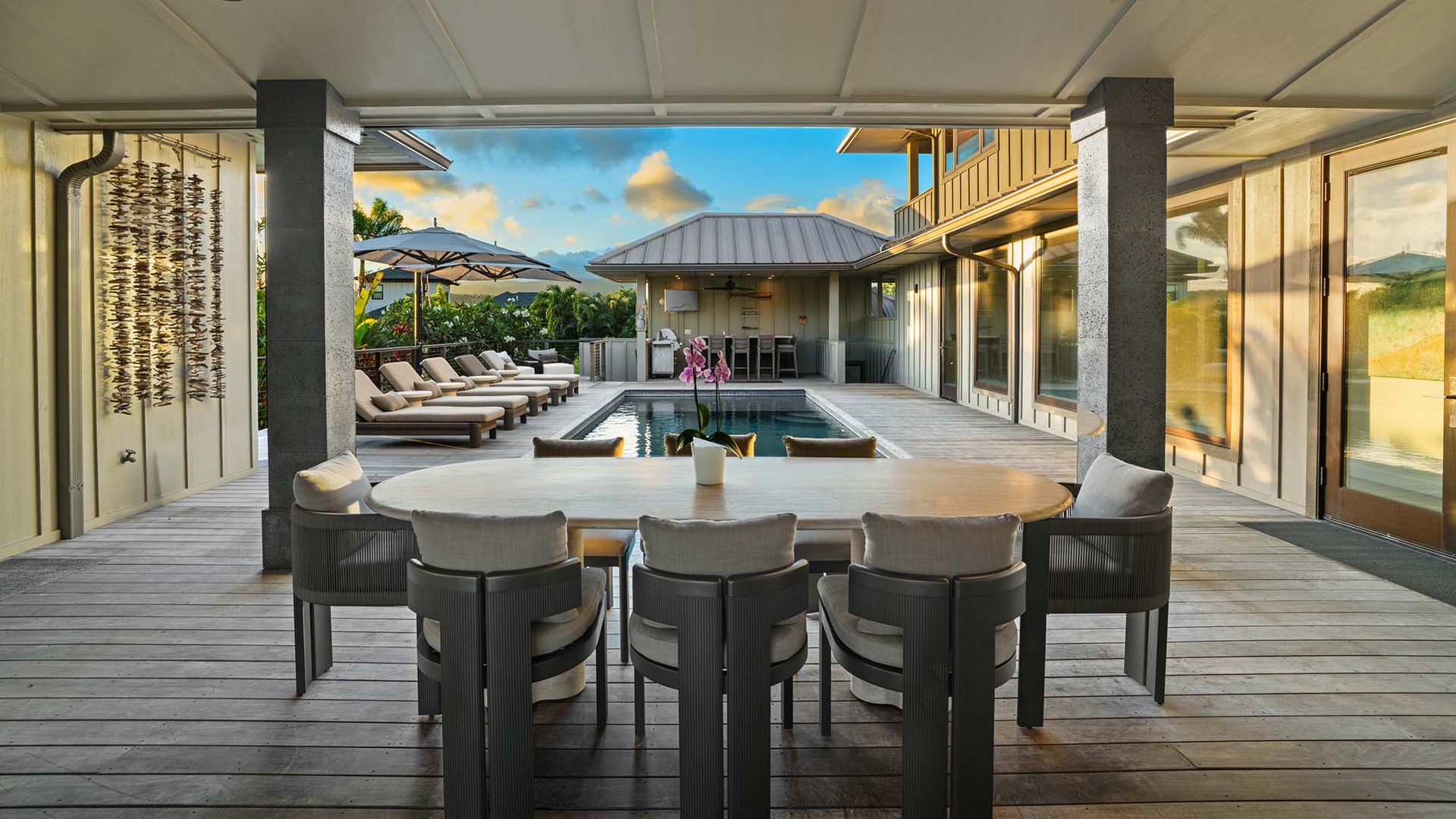 Luxurious covered terrace showcases resort-style living with an elegant dining table, sleek poolside loungers, and a fully-equipped outdoor kitchen framed by tropical surroundings.
