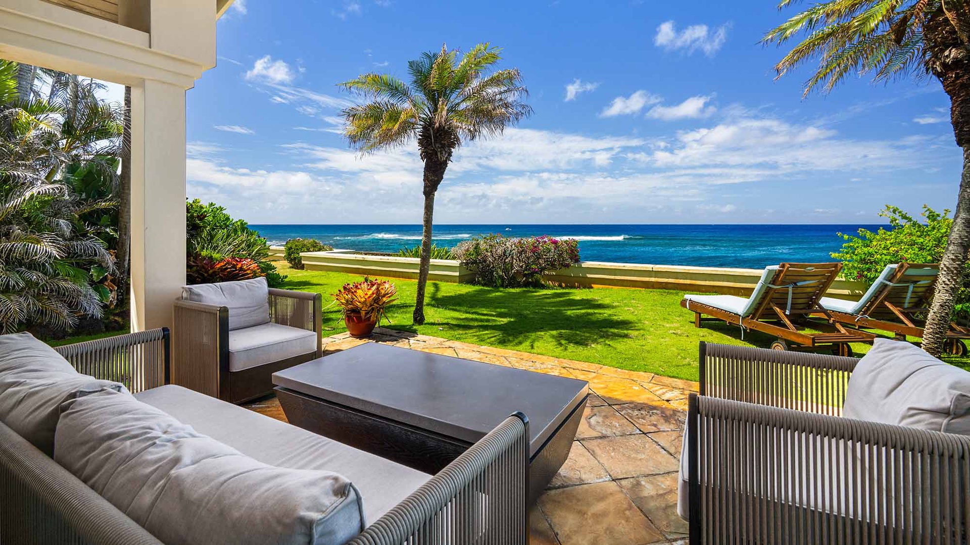 A private coastal setting where vibrant tropical landscaping frames pristine ocean vistas and manicured lawn stretches toward the shore. Palm trees stand sentinel over the beachfront grounds beneath bright island skies.