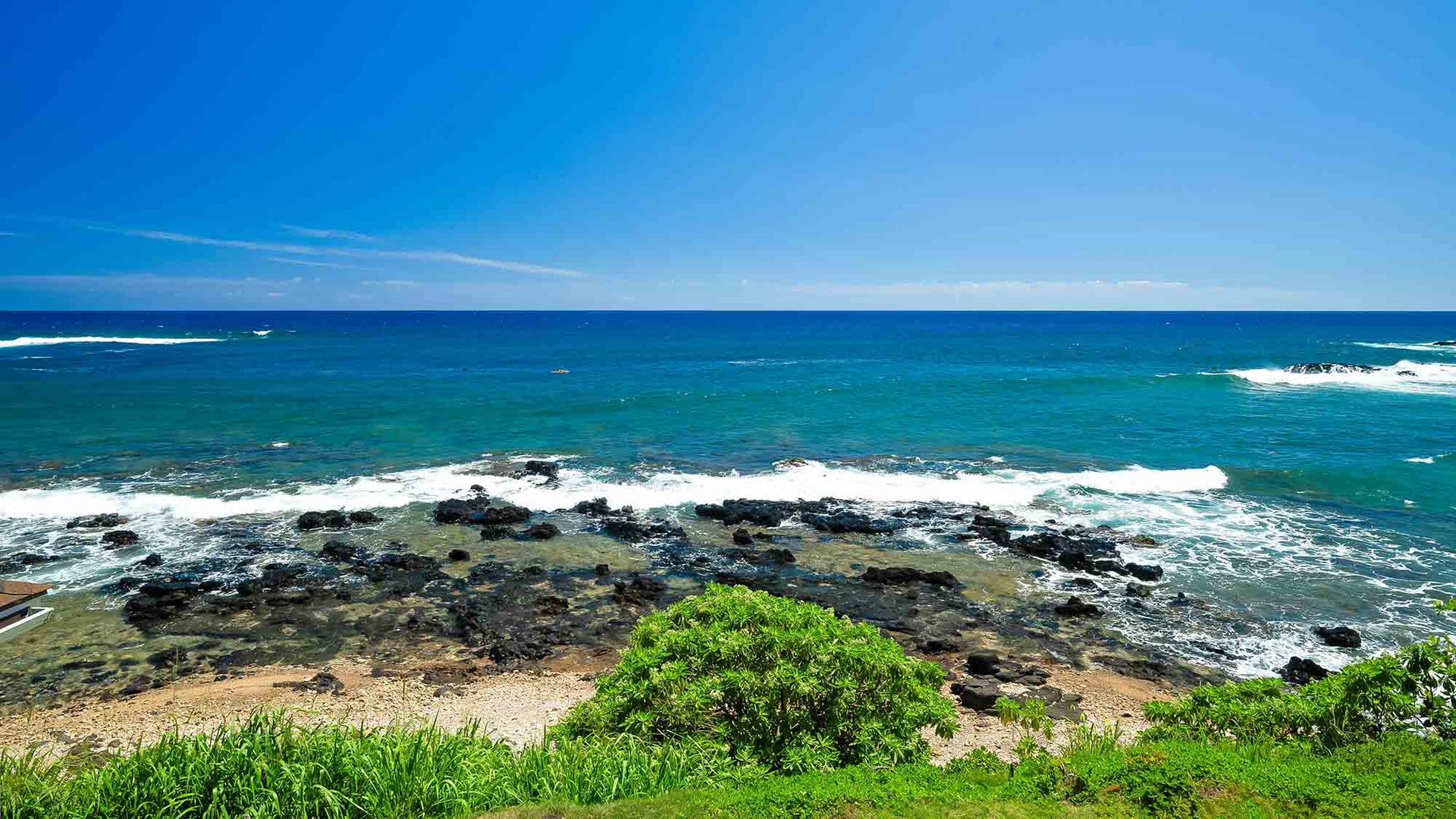 Stunning coastal landscape with brilliant turquoise waters rolling over rocky shores and lush green vegetation under a bright blue sky.