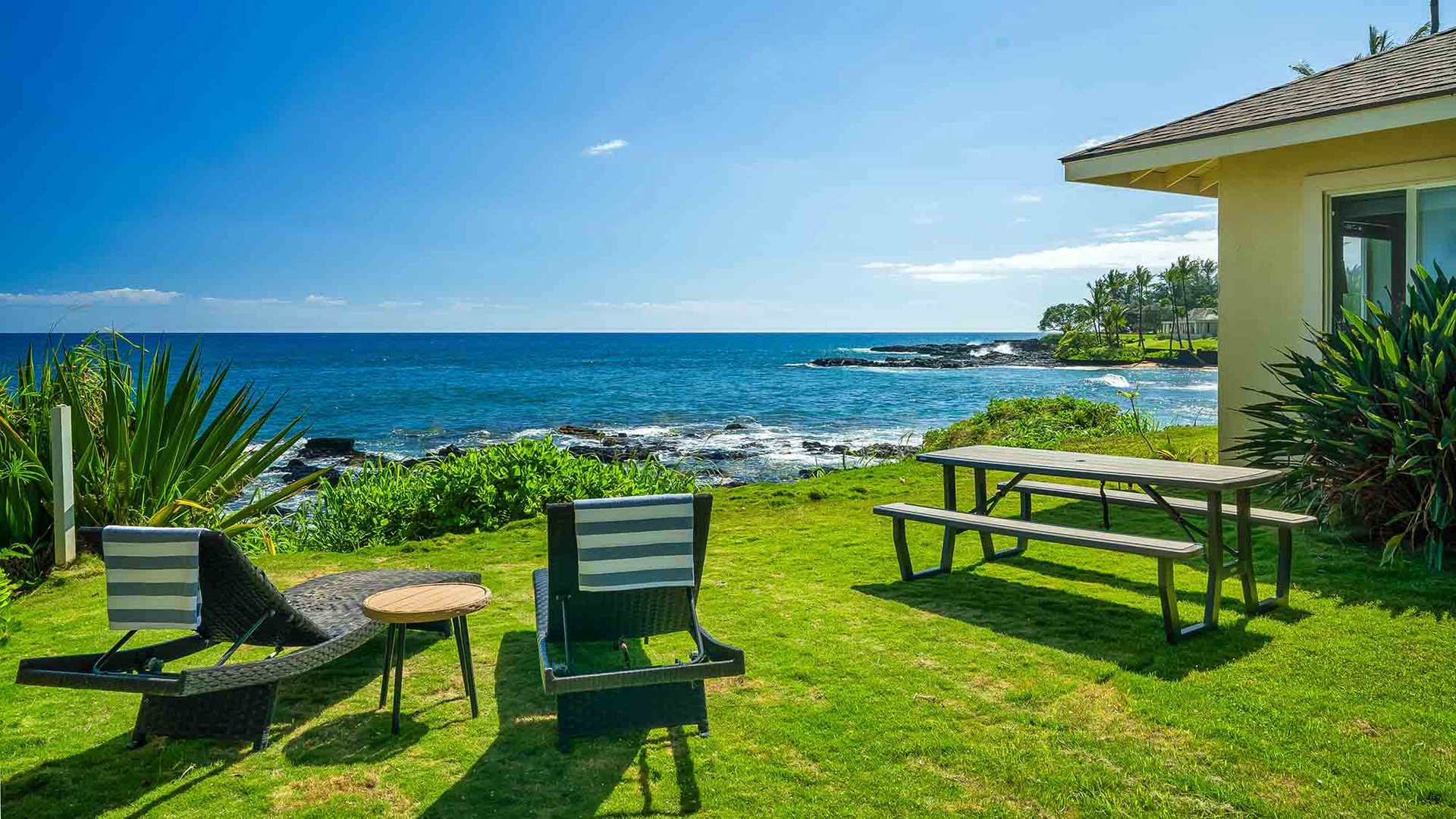 A beachfront property with ocean views, surrounded by tropical greenery and lush lawn. The property is situated along a scenic coastline with volcanic rock formations.
