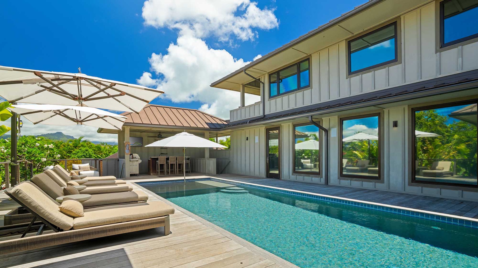 A modern two-story vacation villa featuring a private pool and deck with lounge chairs, surrounded by tropical greenery and mountains under bright blue skies.