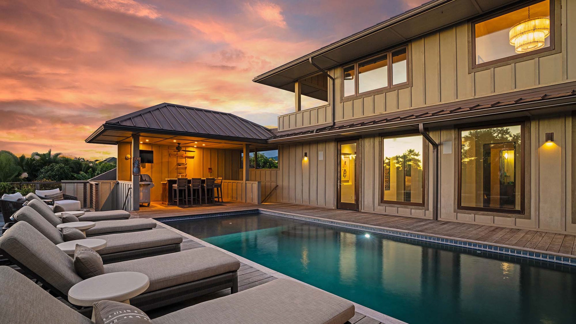 A stunning modern vacation home featuring a private pool, luxurious sun loungers, and covered outdoor entertaining space, all set beneath a breathtaking tropical sunset.