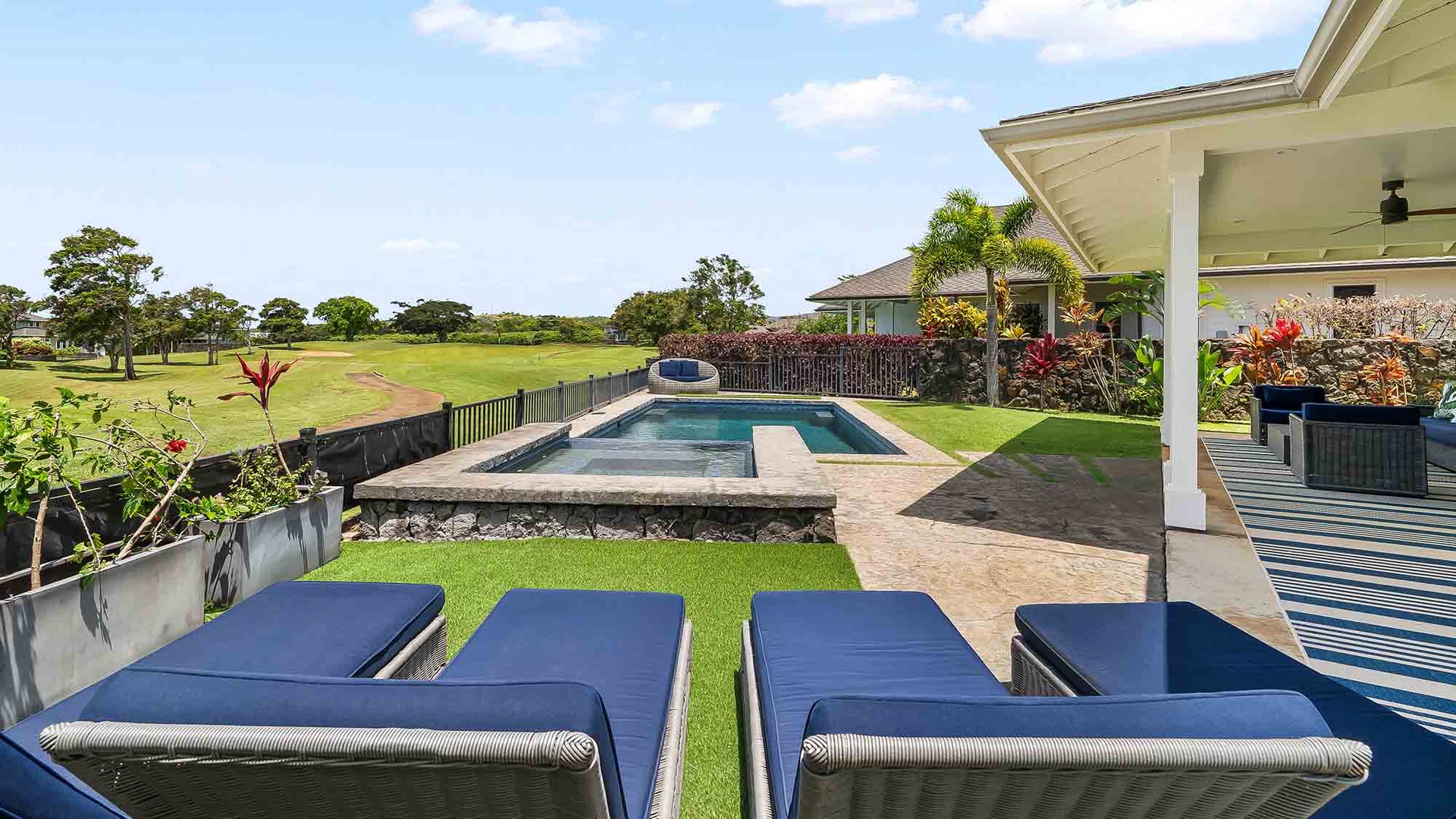 The spacious backyard features a private pool and hot tub surrounded by lush tropical landscaping, with expansive lawn areas and open golf course views. Palm trees and vibrant plantings frame the covered patio area.