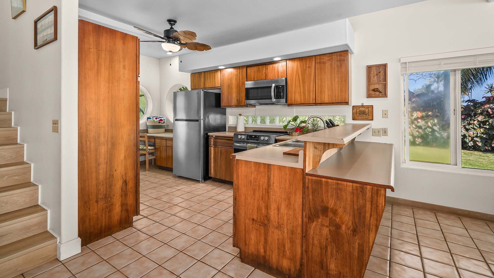 Your fully equipped kitchen features warm wood cabinetry, modern appliances, and tropical breezes from ceiling fans overhead. Natural light pours in through windows framing lush garden views.