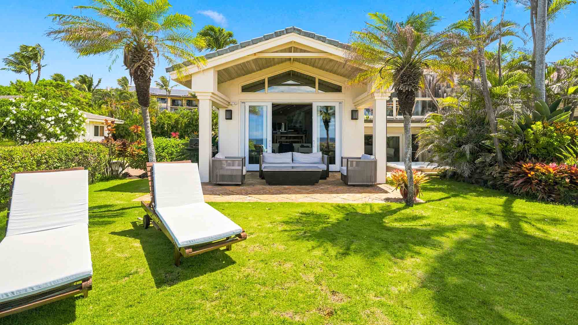 A charming vacation cottage surrounded by lush tropical palms and manicured lawn, featuring a covered patio with seating and two inviting lounge chairs—your perfect retreat in paradise.