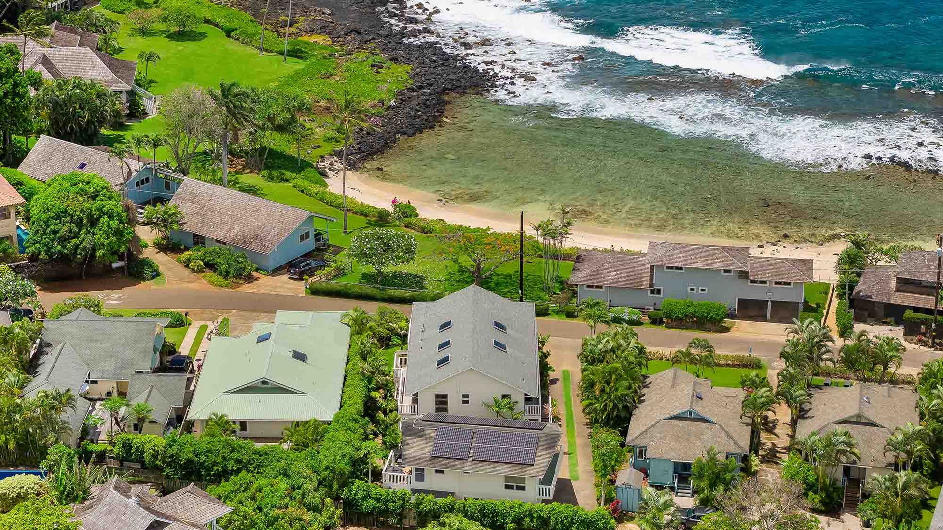 Aerial view of a coastal neighborhood with beachfront vacation homes nestled between lush greenery and turquoise ocean waves, offering serene island living near pristine sandy shores.