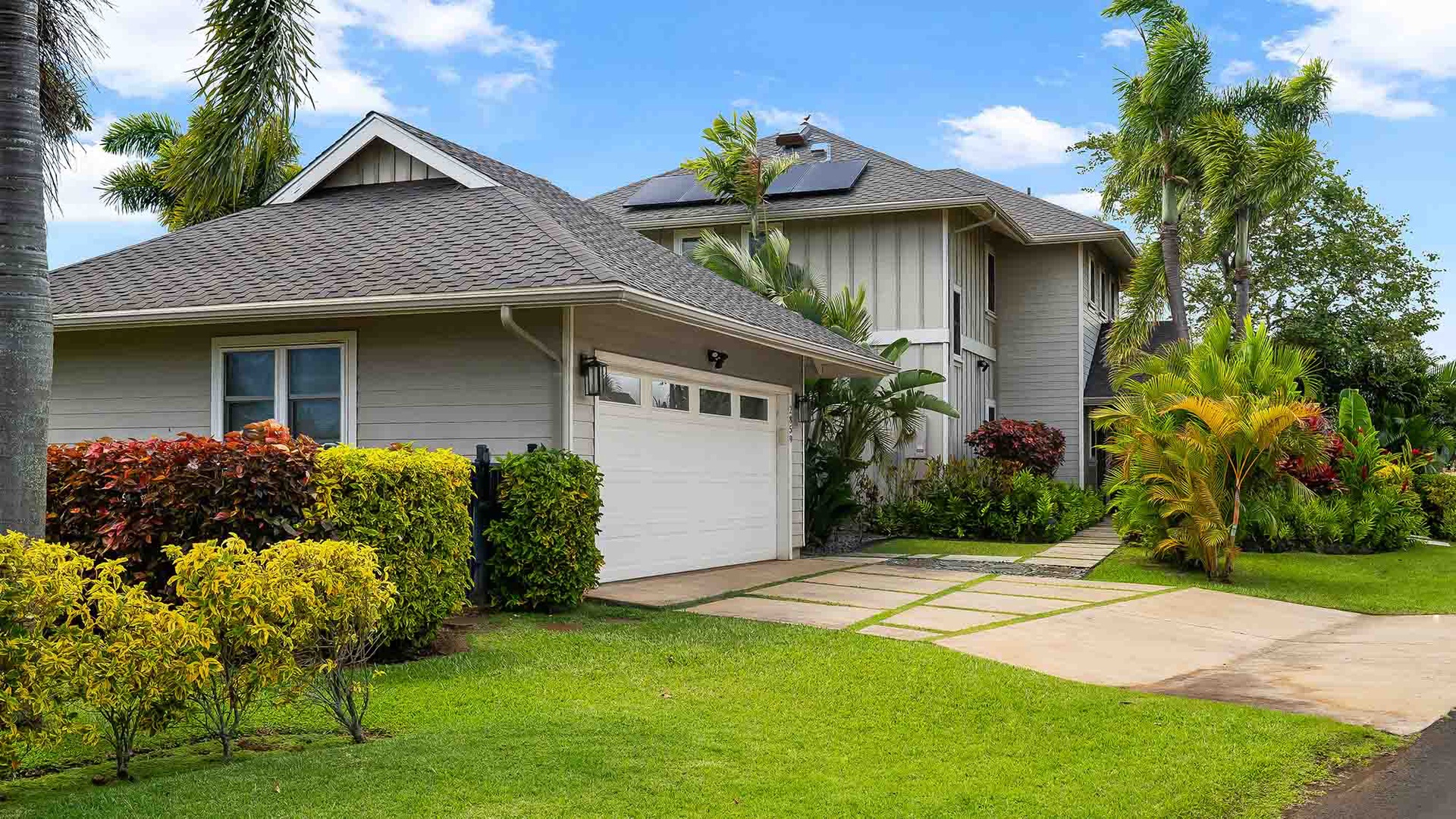 A spacious two-story vacation home with tropical landscaping and lush gardens greets arriving guests. The property features mature palms and colorful plantings surrounding a well-maintained exterior with ample parking.