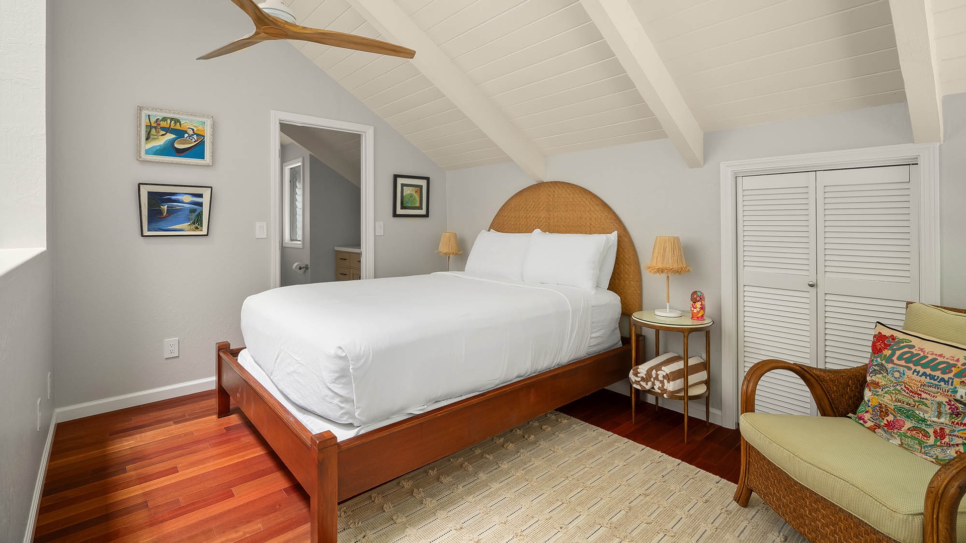 Your peaceful island retreat awaits, with crisp white linens on a stylish rattan-accented bed, warm hardwood floors underfoot, and charming coastal artwork setting a relaxed, tropical vibe.