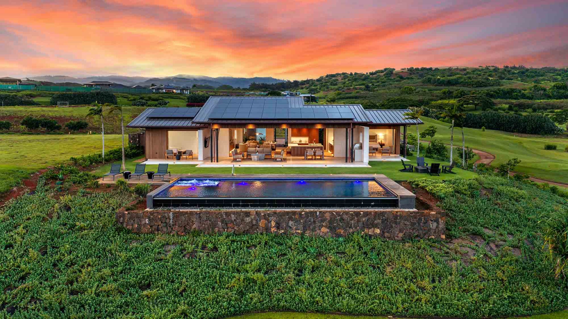 An aerial view captures the property at twilight, showcasing the modern architecture, infinity pool, and surrounding golf course landscape beneath a vibrant sunset sky.