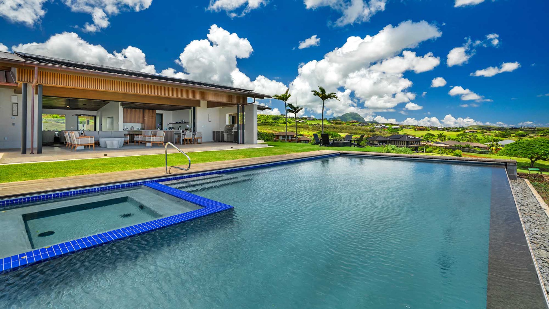 A spacious private pool with sparkling blue tile and an adjacent hot tub awaits, framed by lush green lawn and dramatic mountain vistas under brilliant tropical skies.