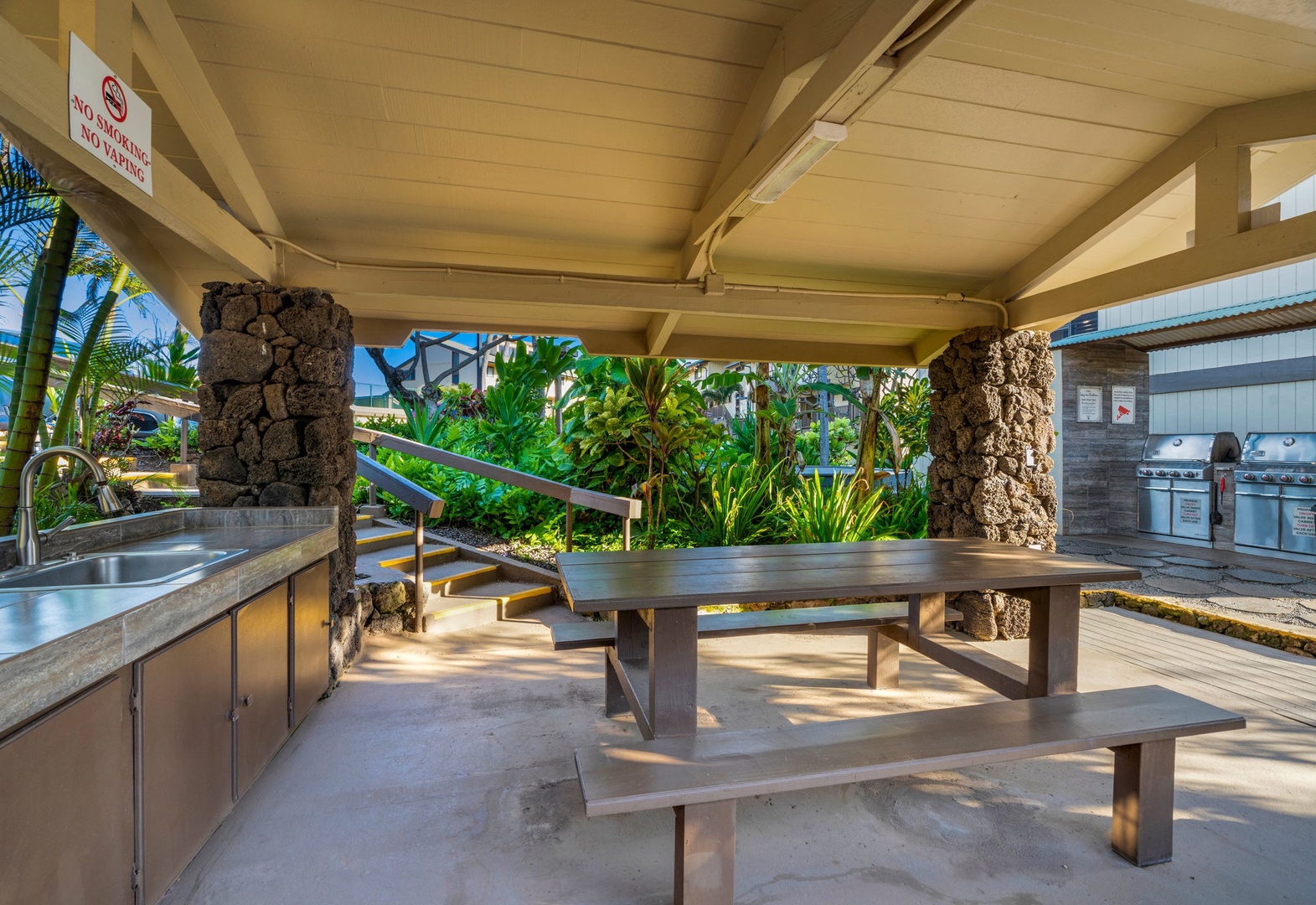 A sheltered outdoor barbecue pavilion features picnic-style seating, stainless steel grills, and a prep sink surrounded by lush tropical landscaping—perfect for gathering with fellow guests.