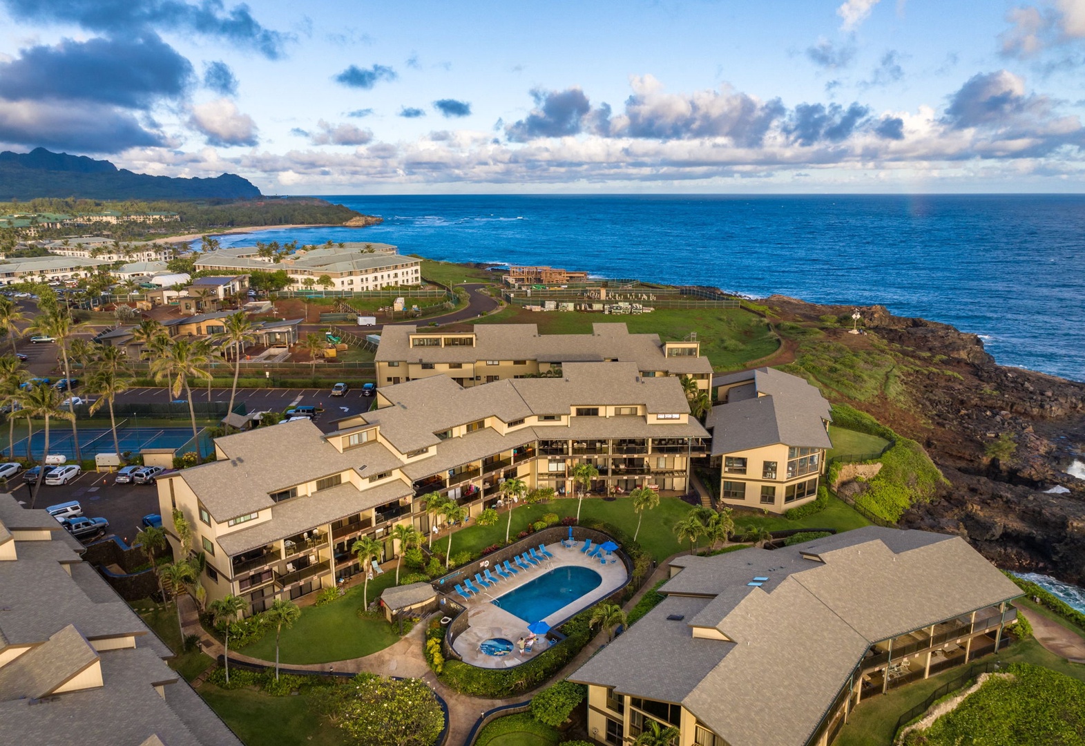 An aerial view captures the property's stunning oceanfront location, nestled between dramatic volcanic coastline and vibrant blue waters, with mountains framing the resort community.