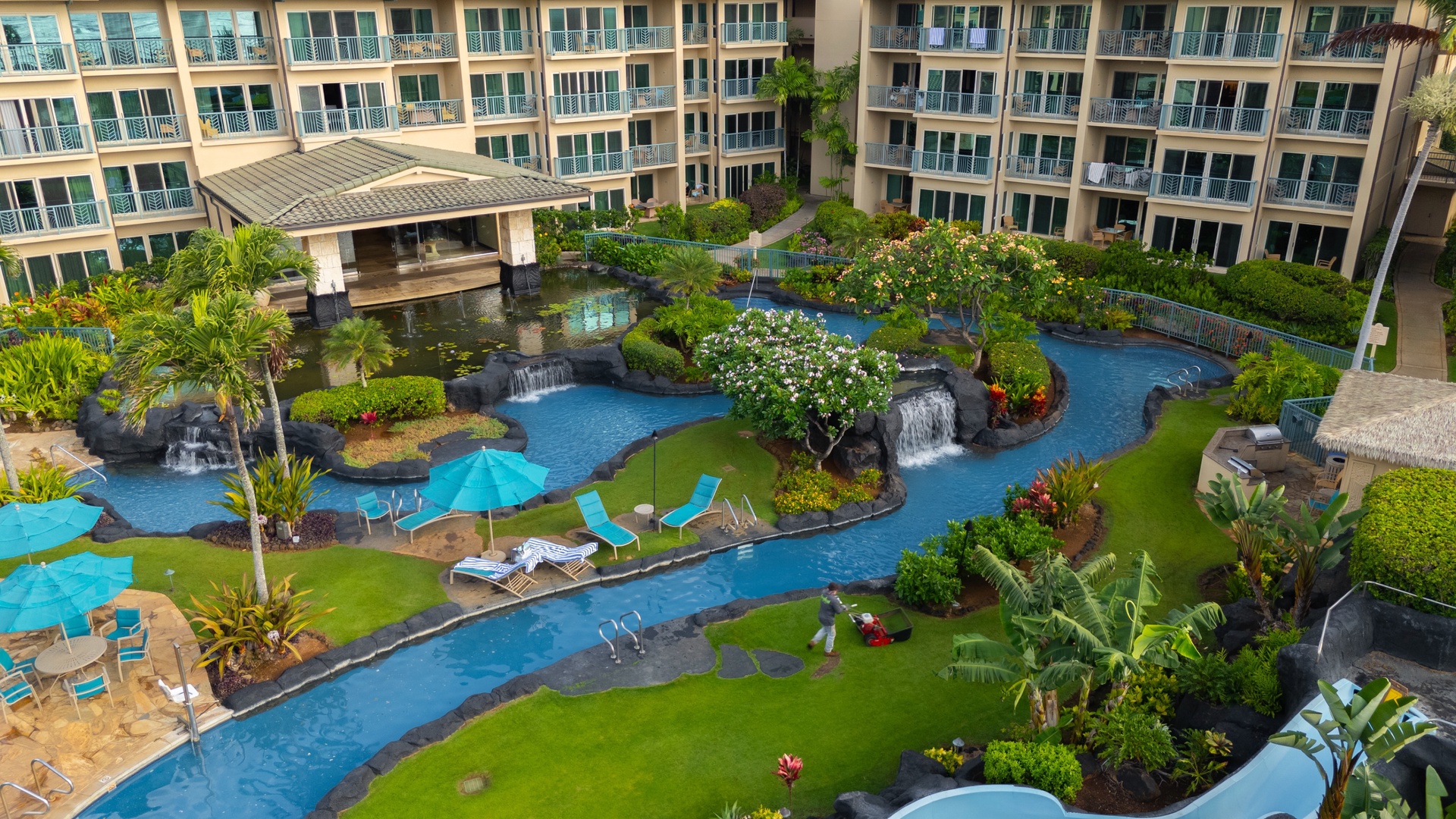 An aerial view of the property reveals a lush tropical oasis with meandering pools, cascading waterfalls, and vibrant landscaping nestled between multi-story buildings with private balconies.