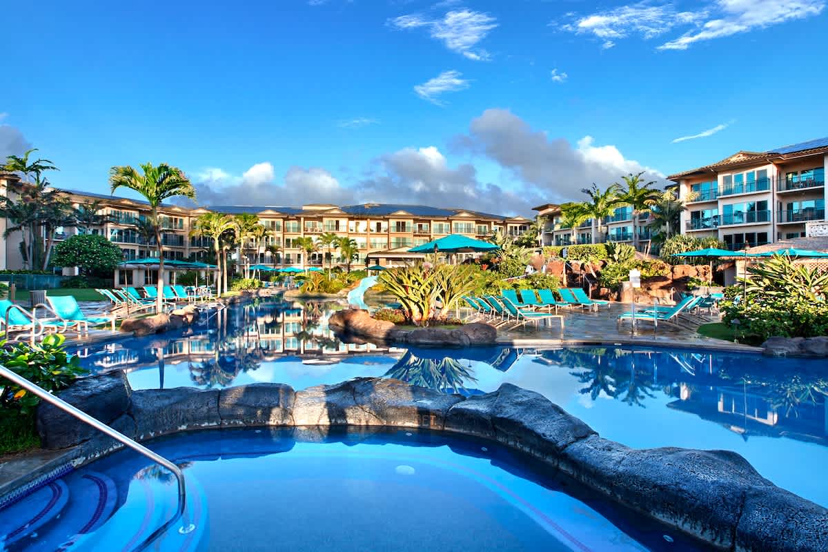 A multi-level resort property featuring a tropical lagoon-style pool area with waterfalls, lava rock accents, and multiple sections for relaxation surrounded by lush palms and turquoise lounge chairs.