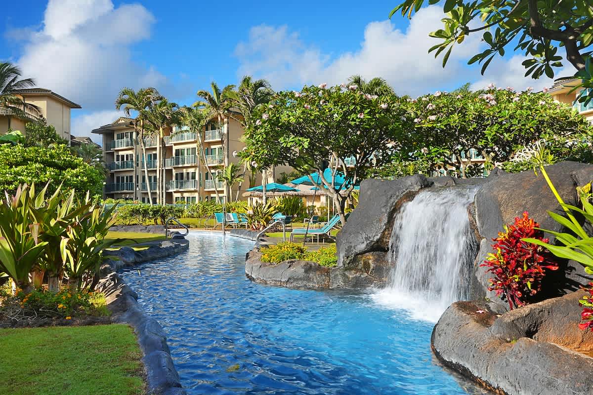A tropical oasis set within lush landscaping with a tranquil waterfall feature, vibrant flowering plants, and inviting turquoise waters framed by verdant greenery and palms.