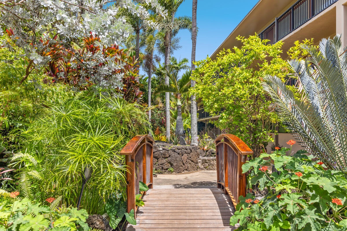 A wooden walkway bridges tropical gardens with vibrant palms, flowering plants, and stone features leading to the building.