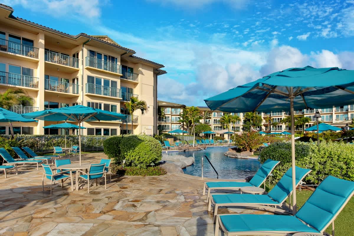 The property features a luxurious outdoor pool surrounded by vibrant turquoise loungers and umbrellas, set against a stunning multi-story building with private balconies and lush tropical landscaping under bright blue skies.