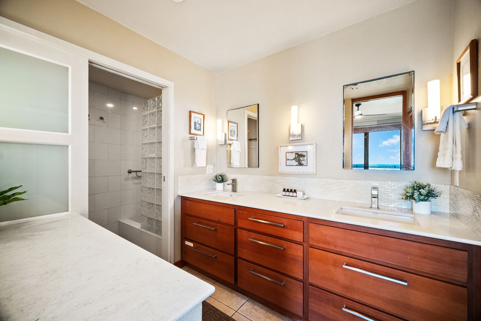 Step into your elegant bathroom featuring dual sinks, rich wood cabinetry, and a spacious walk-in shower. Natural light pours through the window while you freshen up, framing stunning ocean views to start your day right.