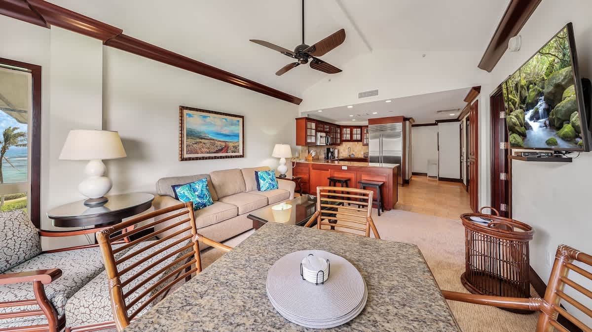 Relax in your open-concept living area where rich wood accents and tropical decor create a warm island vibe. Unwind on the plush sofa, catch ocean views through the doors, and enjoy the fully equipped kitchen with modern appliances.