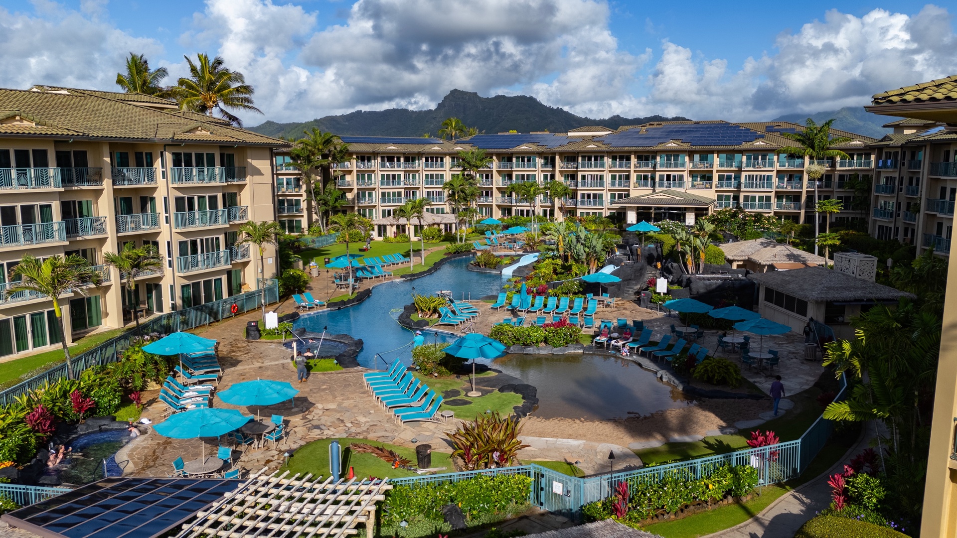 A resort-style property surrounded by tropical landscaping with mountain views, featuring multiple buildings arranged around a central pool complex with vibrant blue umbrellas and lush greenery under dramatic skies.