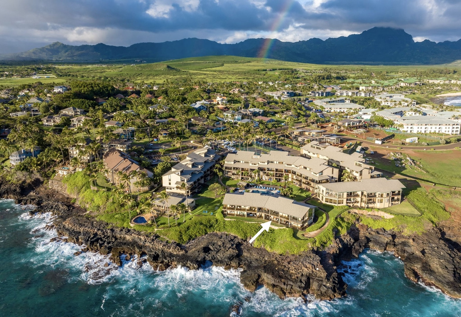 Aerial view of the resort property perched on dramatic volcanic coastline, surrounded by lush tropical landscapes and backed by majestic mountain ranges under vibrant skies.
