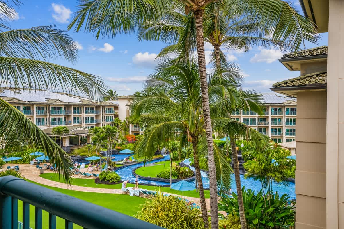 The property features a sprawling pool complex surrounded by lush tropical landscaping and palm trees, with multi-story buildings offering balcony views and abundant green space throughout the grounds.