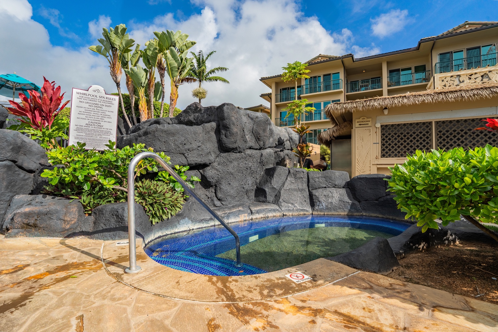 A tropical property featuring volcanic rock accents, lush landscaping with palm trees, and blue-tiled spa pools set against multi-story resort buildings.