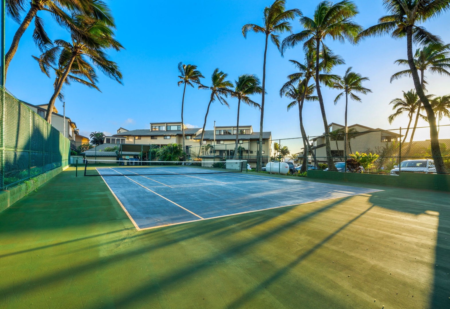 A pristine tennis court surrounded by swaying palm trees offers a perfect setting for an active tropical getaway, with the property buildings just steps away.