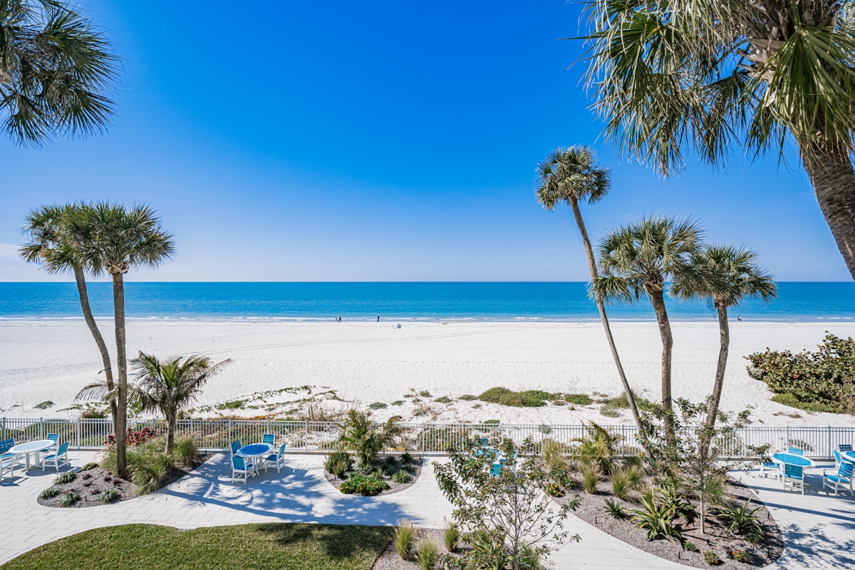 Stunning Gulf Coast beachfront with white sand stretching before you, swaying palms, and turquoise waters. The landscaped outdoor patio offers a perfect spot to soak up the sun and sea breeze.