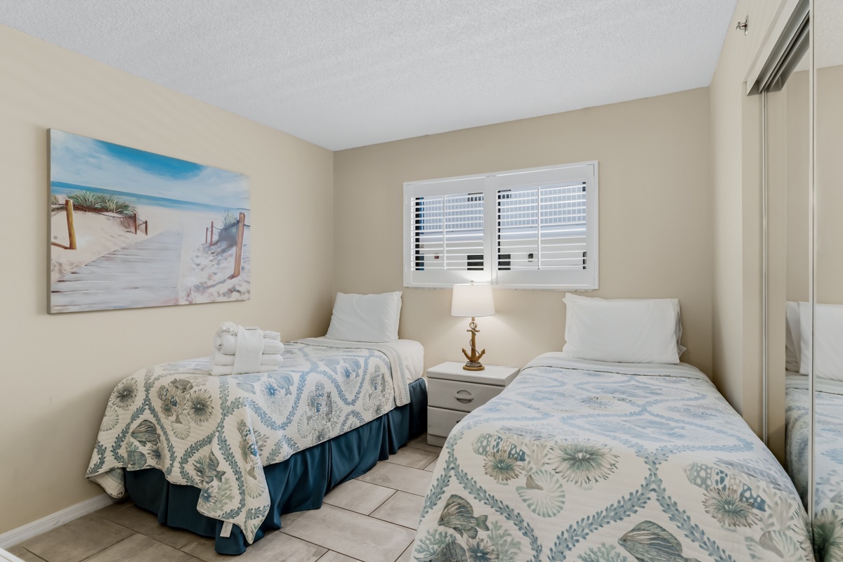Relax in this peaceful twin bedroom with coastal charm. Your comfortable beds await, dressed in soft blue and white linens that echo the serene beach artwork above.