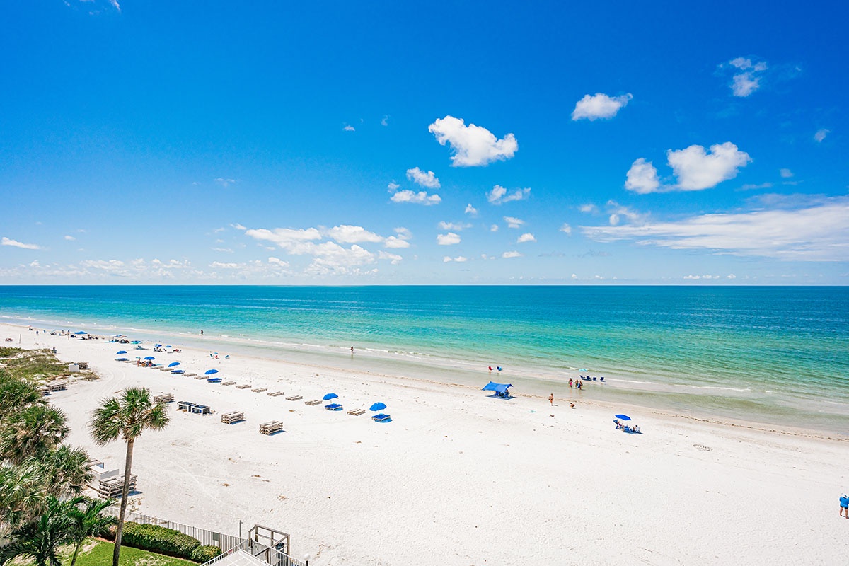 Pristine white sand beach stretches along turquoise waters, dotted with colorful umbrellas and palm trees under brilliant blue skies.