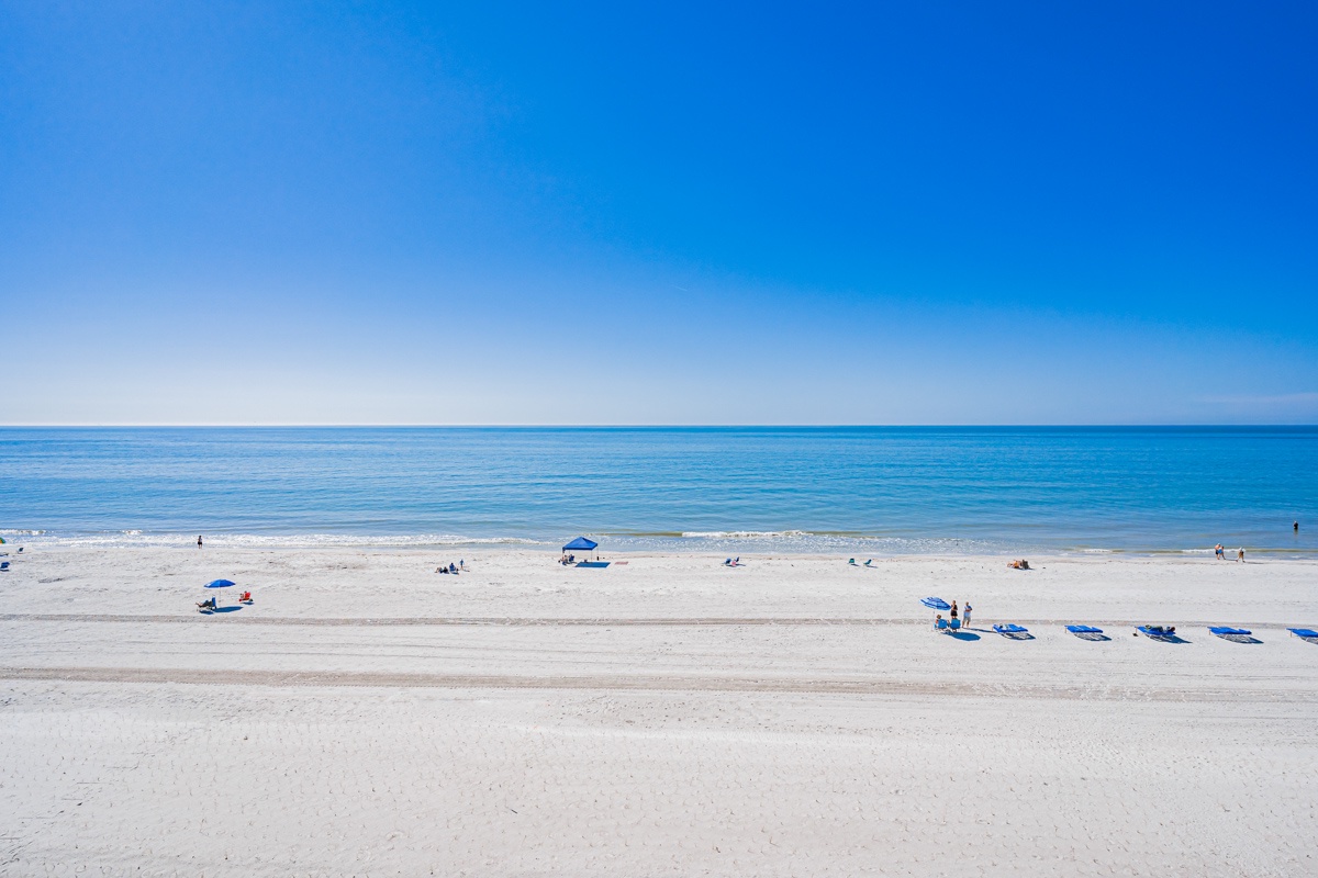 Pristine white sand beach stretches along crystal-clear waters under brilliant blue skies, with convenient beach umbrellas and loungers available.