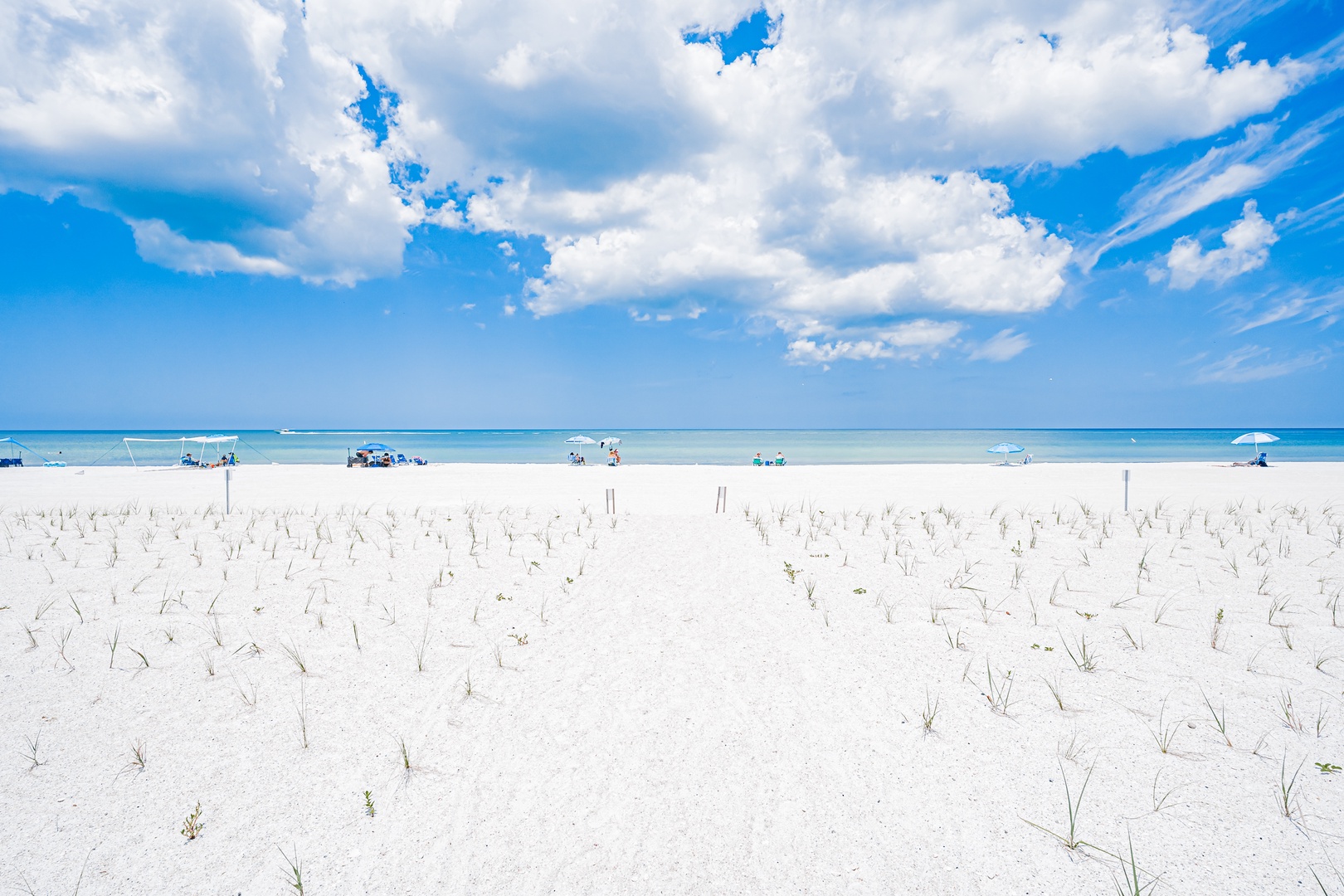 Powder-soft white sand stretches beneath endless blue skies, meeting calm turquoise waters where guests unwind under umbrellas, creating an idyllic beachside retreat.