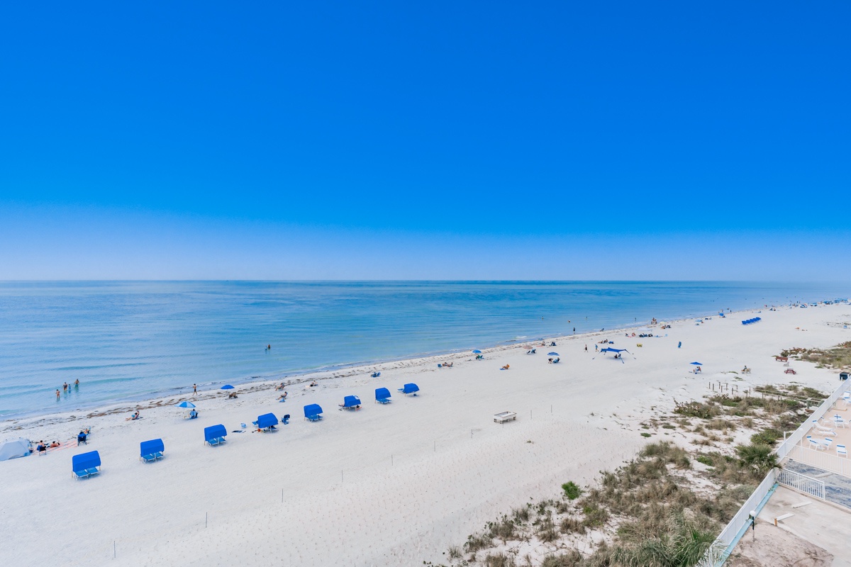 Pristine white sand beach stretches along crystal-clear waters, with blue umbrellas dotting the shoreline and gentle waves lapping the coast.