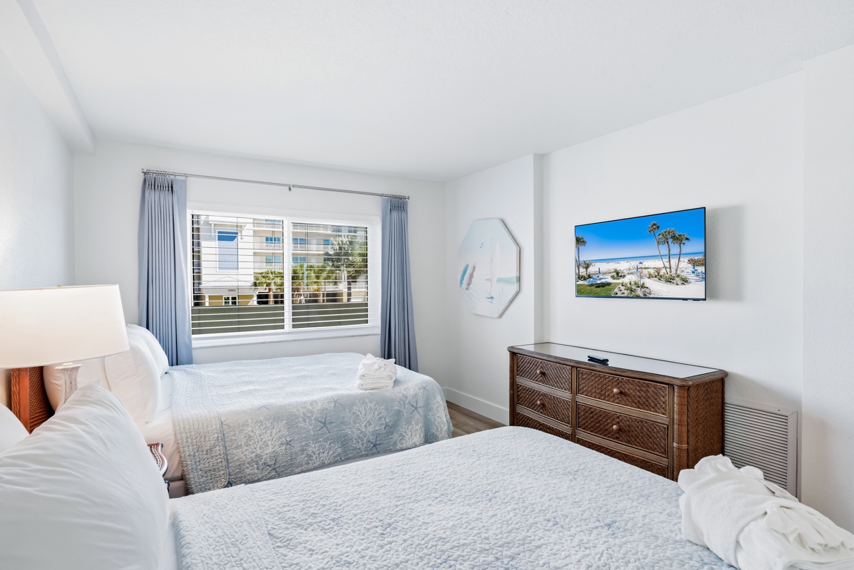 Rest peacefully in your bright bedroom with two comfortable beds, tropical artwork, and blue curtains framing city views.