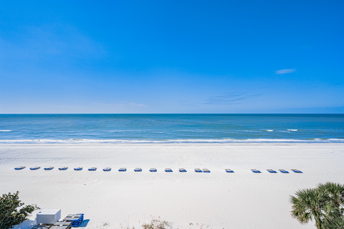 Pristine white sand beach stretches endlessly with turquoise waters and blue beach chairs dotting the shoreline under clear skies.