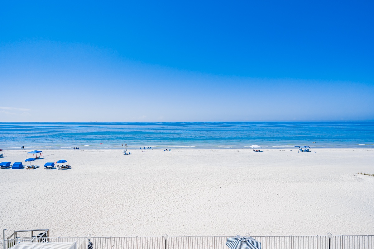 A pristine white-sand beach stretches beneath an endless blue sky, where gentle waves roll in and beachgoers enjoy the sunshine. Your perfect beach day awaits just steps from your door.