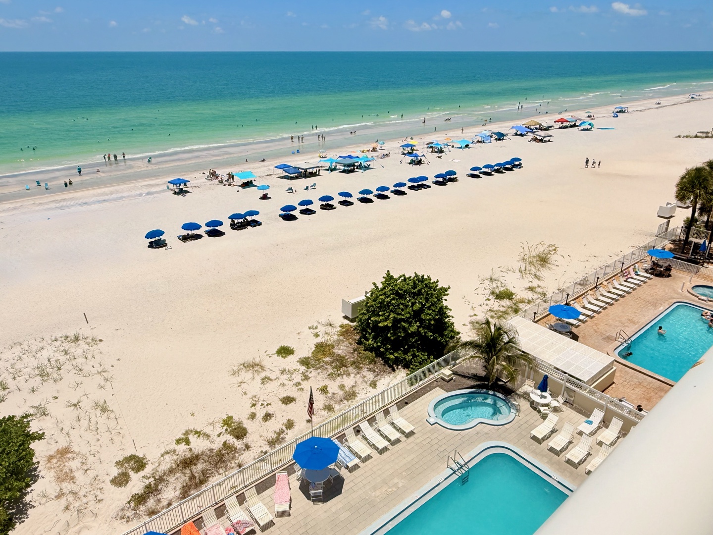 An aerial view of the beachfront property reveals pristine white sand, turquoise Gulf waters, and two sparkling pools with a hot tub, all steps from rows of complimentary beach umbrellas and loungers.