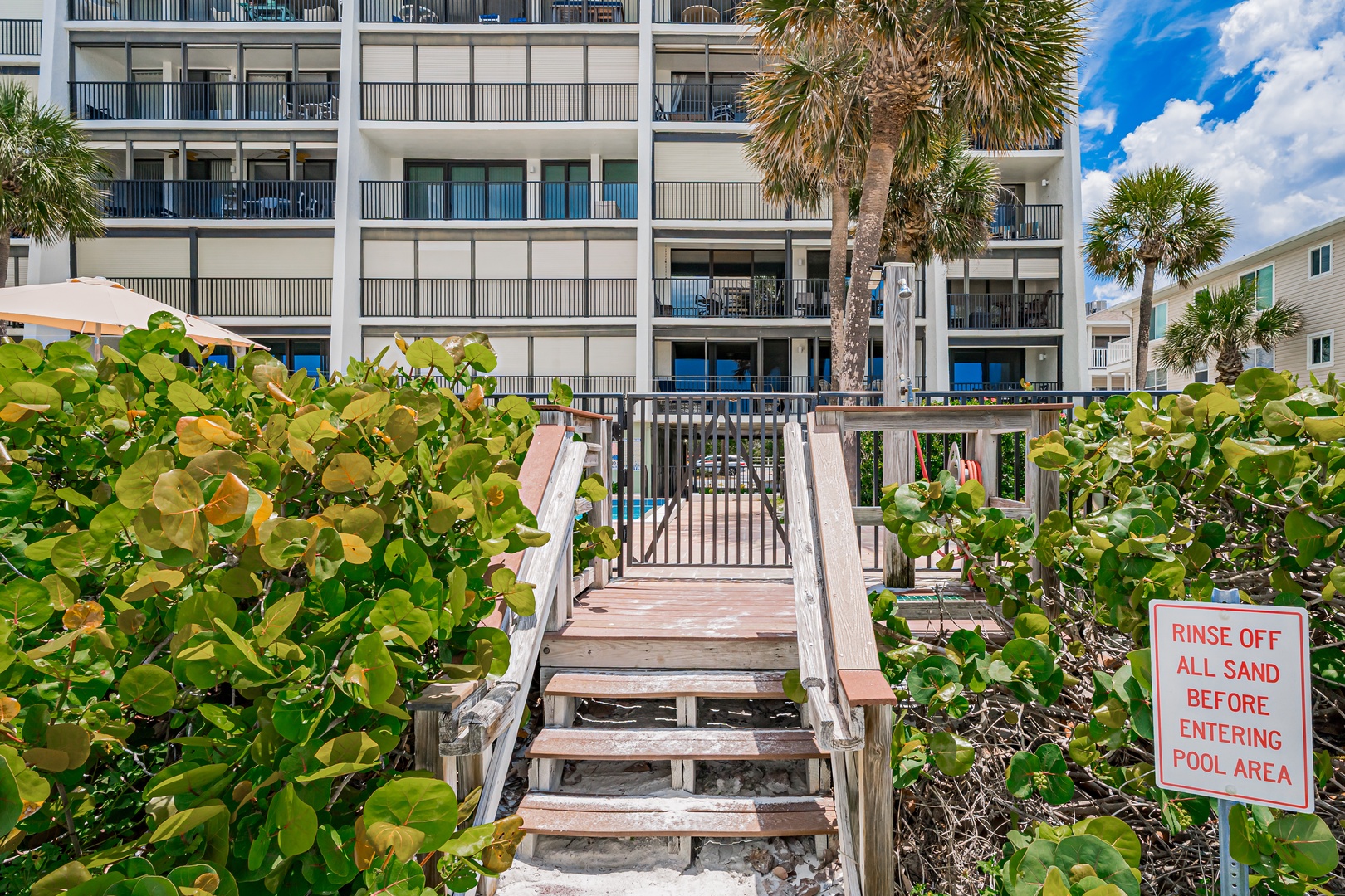 Beachfront property with direct beach access via wooden walkway, featuring private balconies, lush tropical landscaping with palm trees, and ocean views along the Florida coastline.